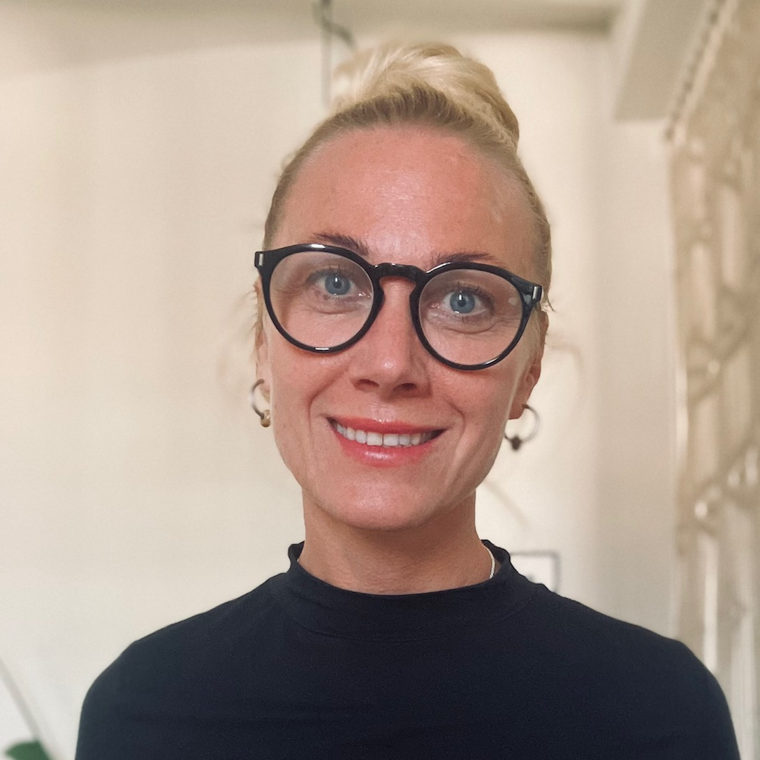 En la foto de perfil, Linn Biorklund se ve de frente, sonriendo. Mujer blanca, con gafas de lectura negras redondas, pelo rubio atado en el topo de la cabeza en forma de coque. Lleva una camiseta negra de cuello cerrado y pendientes de aro dorados.